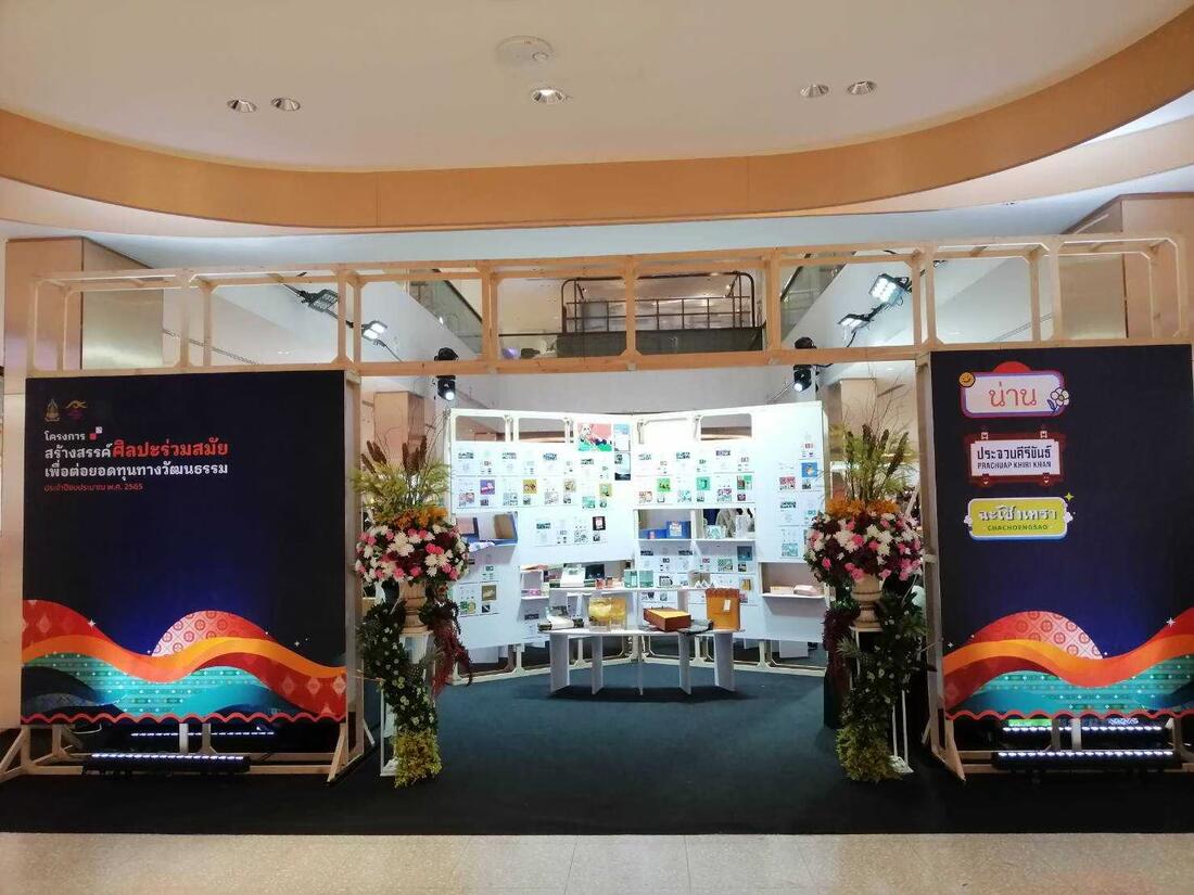 บูธนิทรรศการในห้างสรรพสินค้า มีชั้นวางจัดแสดงผลงานและแจกันดอกไม้สองข้าง